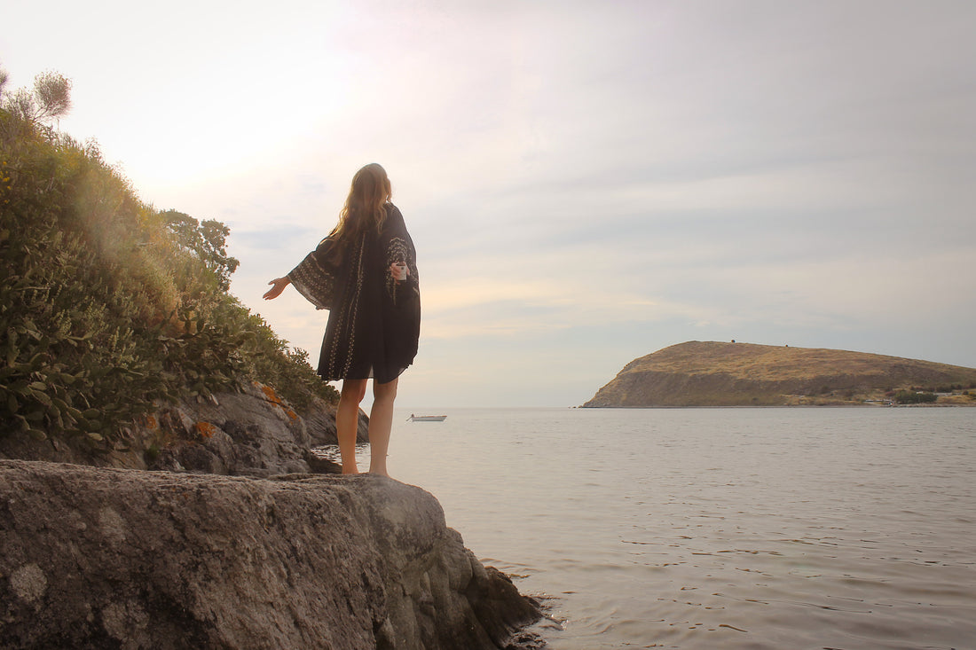 Kvinne står på en klippe og ser utover havet mens hun holder en flaske magnesiumolje med armene opp og litt bak